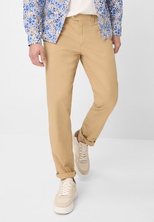 Chino in cotone beige con vestibilità dritta, dotati di orli arrotolati, abbinati a sneakers di colore chiaro. La camicia presenta un motivo floreale blu.