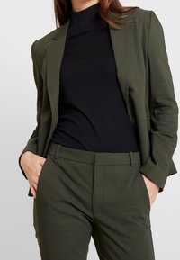 Olivově zelený blazer přes černý žebrovaný rolák, s upravenými kalhotami. Má úzký střih, zaoblené klopy a boční kapsy.