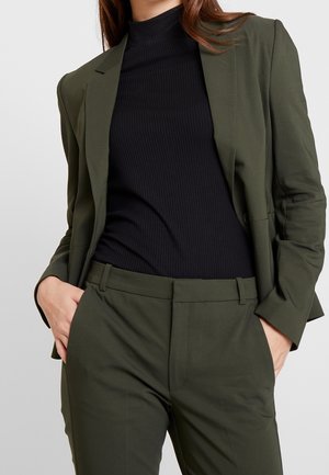 Blazer verde oliva sobre un cuello de tortuga negro con canalé, combinado con pantalones a medida. Presenta un corte ajustado, solapas de muesca y bolsillos laterales.
