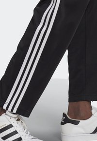 Pantaloni sportivi neri con tre strisce verticali bianche sui lati, abbinati a sneaker bianche con dettagli neri e design classico.