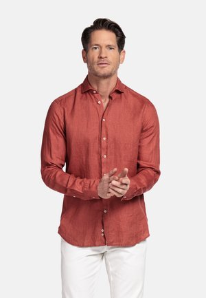 Man met een roestkleurig overhemd met lange mouwen en witte broek, staand tegen een effen lichte achtergrond, met gevouwen handen.