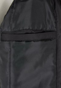 Tela de chaqueta acolchada negra con un bolsillo en la costura lateral y una solapa oculta. Textura suave con detalles de costura a lo largo de los bordes.