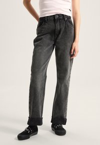 Jeans negros de tiro alto con un acabado ligeramente descolorido, piernas anchas, puños deshilachados y diseño clásico de cinco bolsillos. Combinados con zapatillas negras.