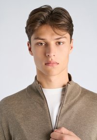 Jeune homme aux cheveux bruns courts fermant la fermeture éclair d'un pull beige par-dessus une chemise blanche, face à l'avant sur un fond uni.