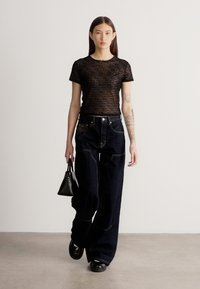 Νέα γυναίκα με μακριά σκούρα μαλλιά φοράει διαφανές μαύρο ριγέ τοπ, φαρδιά σκούρα τζιν, μαύρα παπούτσια και κρατά μια μικρή μαύρη τσάντα.