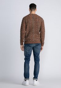 Brown cable-knit sweater with red accents, long sleeves, ribbed hem, worn with blue jeans and white sneakers, shown from the back.