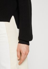 Czarny dziergany sweter z prążkowanymi mankietami, z unikalnym ukośnym dołem. Ręka oparta na jasnym, dopasowanym ubraniu.