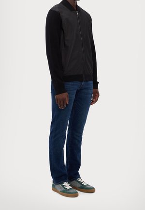 Homme portant une veste noire zippée, un jean bleu foncé et des baskets vertes et grises avec des lacets blancs, debout sur un fond blanc.