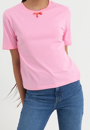 Vrouw draagt een lichtroze shirt met korte mouwen, een kleine rode strik bij de halslijn en een blauwe spijkerbroek, tegen een effen achtergrond.