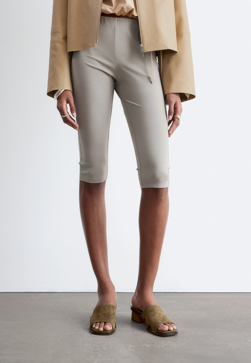 Personne portant des leggings courts beige, une veste courte beige et des mules à talons ouvertes kaki, debout sur un sol gris.