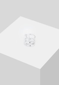 Anello in metallo argento con tre anelli impilati, ciascuno dalla forma liscia e arrotondata con una finitura lucida, esposto su una superficie bianca.