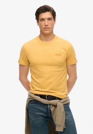 Gul bomulds t-shirt med korte ærmer, rund hals og lille broderet logo. Båret med blå jeans og en beige sweater knyttet om livet.