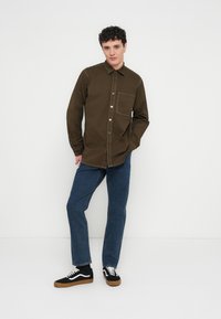 Ung man med lockigt hår klädd i brun skjorta med knappar, blå jeans och svarta sneakers, stående mot en vit bakgrund.