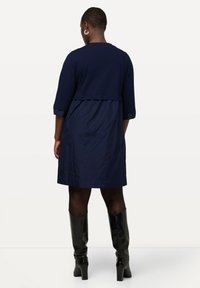 Dunkelblaues Kleid mit Dreiviertelärmeln, ausgestelltem Saum und geschichtetem Design. Getragen mit kniehohen schwarzen Stiefeln. Glatte Stofftextur.