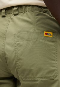 Žalios spalvos Fjällräven kelnės iš arti, rodantis galinę kišenę ir geltoną logotipo lopelį ant kišenės krašto.