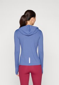 Långärmad tränings-topp i blått med huva, tillverkad av ett slätt, elastiskt material. Har en vit detalj på den nedre ryggen. Matchas med vinröda leggings.