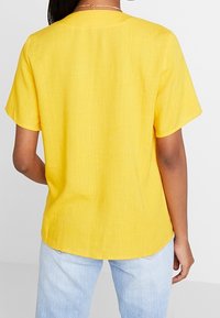 Krátký rukáv v jasně žluté barvě, s kulatým výstřihem a texturovanou tkaninou. Pohled zezadu ukazuje rovný lem.