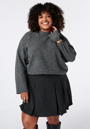Lachende vrouw met krullend haar, gekleed in een grijze trui, donkere geplooide rok, gouden armbanden en zwarte laarzen tegen een effen achtergrond.