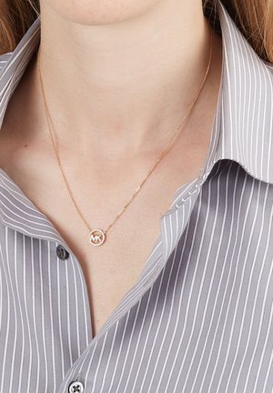 Collana d'oro con pendente circolare con le lettere "MK", indossata sopra una camicia a righe grigio chiaro e bianca, che mostra un delicato design della catena.
