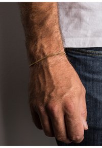 Gold chain bracelet on a wrist with visible veins and hair; hand is relaxed, wearing blue denim jeans, neutral background.