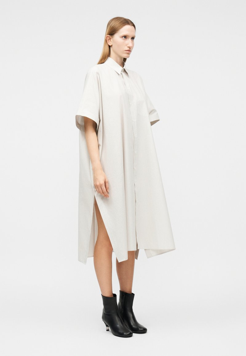 Femme debout portant une robe midi beige clair, ample, boutonnée, avec des fentes latérales et des bottines noires, sur un fond uni.