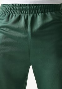 Πράσινο αθλητικό παντελόνι κατασκευασμένο από λεία, γυαλιστερή ύλη. Διαθέτει ελαστική μέση με ρυτιδωμένη λεπτομέρεια και ίσια γραμμή.
