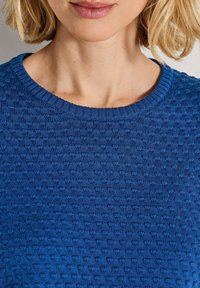 Nahaufnahme des Halses und der Lippen einer Frau, die einen strukturierten blauen Strickpullover mit rundem Ausschnitt trägt.