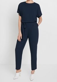 Mörkblå byxdress med korta ärmar, midjeband, avslappnad passform, avsmalnande ben och slät tygkvalitet. Bärs med vita klackar.