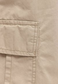 Gros plan sur un tissu beige avec une poche plaquée rectangulaire et des détails de couture apparents.