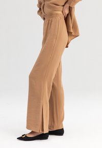 Pantaloni a gamba larga color marrone chiaro, realizzati in tessuto leggero, con spacchi laterali all'orlo. Abbinati a ballerine nere a punta con una fibbia dorata.