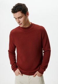 Czerwony sweter o fakturze z okrągłym dekoltem i długimi rękawami, z prążkowanym dołem i mankietami, noszony na jasnych spodniach.