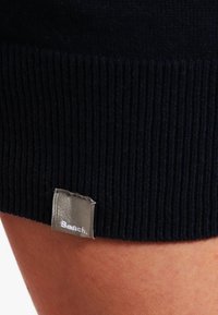 Marinblå ribbad trikå med en silveretikett som visar "Bench." vid nederkanten. Har en åtsittande passform och mjuk textur.