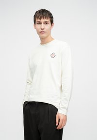 Hosszú ujjú, krémszínű pamut ing, kerek nyakkivágással és egy kis nyomtatott logóval a mellkasán. Fekete nadrággal párosítva.