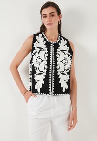 Zwarte vest met witte bloemenborduursels en kralendetails langs de randen. Mouwloos ontwerp, gecombineerd met lichte broeken.