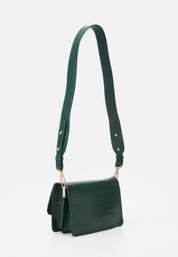 Grön väskor i syntetiskt krokodilskinn med en lång justerbar rem, som visar en snygg, modern design mot en vit bakgrund.
