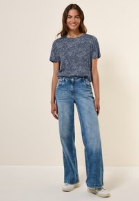 T-shirt à manches courtes bleu marine avec un motif de lignes ondulées blanches, associé à un jean large bleu clair et des baskets blanches.