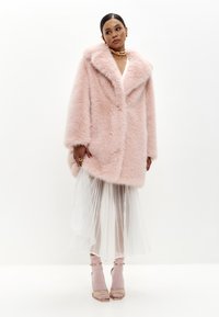 Faux fur frakke i blød lyserød, oversized design med stor krave, parret med en hvid plissee-skirt og lyserøde hæle-sandaler. Guld accenter.