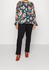 Blomstret bluse med sort baggrund, lange gennemsigtige ærmer og rund halsudskæring, parret med sorte tætsiddende bukser og brune hæle.