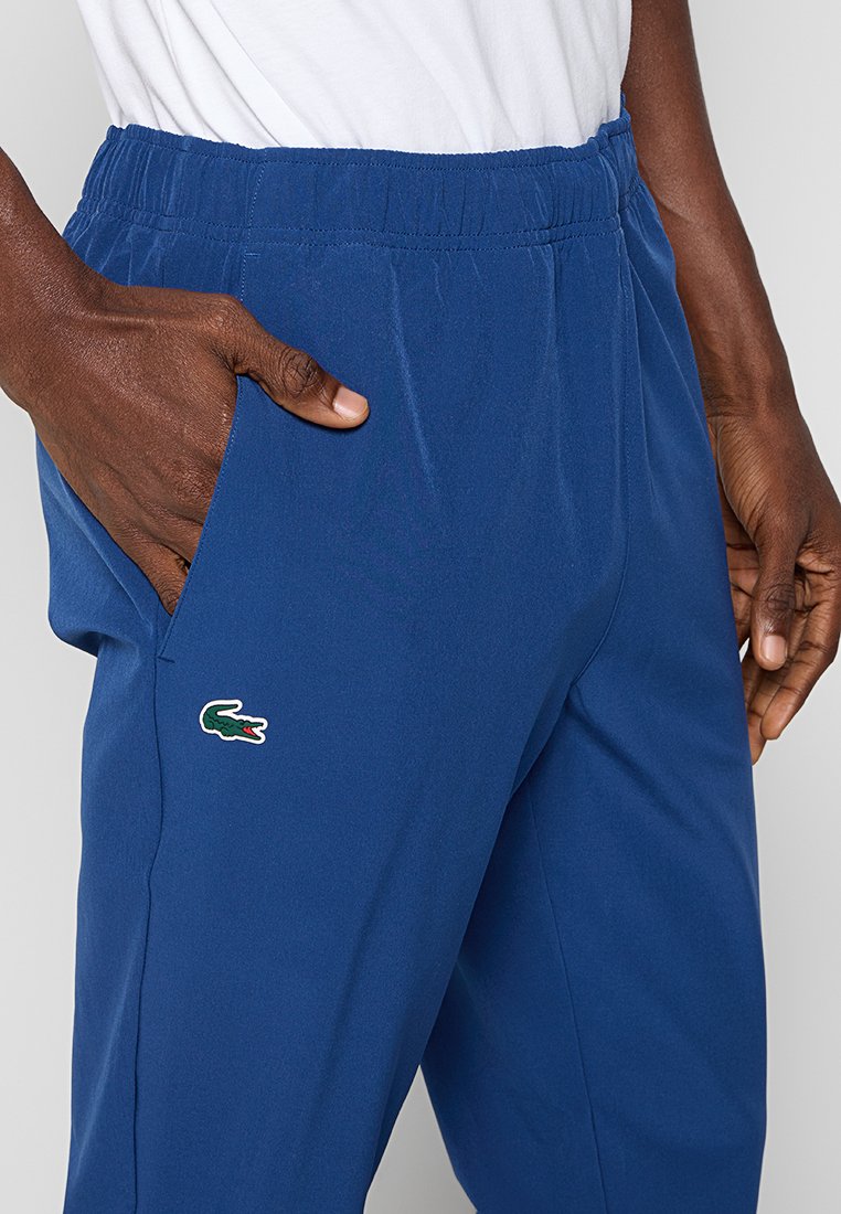 Persona di pelle scura che indossa pantaloni blu con vita elastica e un logo di coccodrillo verde, insieme a una camicia bianca, con una mano parzialmente in tasca.