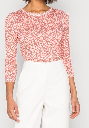 Femme portant une chemise à manches longues ajustée avec un petit imprimé floral rouge et un pantalon blanc taille haute, debout devant un fond uni.