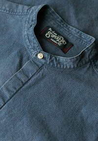 Blauwe gestructureerde shirt met een bandkraag, voorzien van een knoopsluiting en een merklabel. Het materiaal heeft een subtiele ruitpatroon.