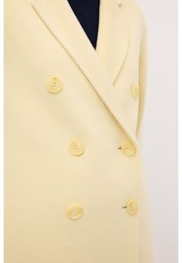 Abrigo amarillo de doble botonadura confeccionado en una tela suave, con seis botones a juego y solapas muesca, que presenta una textura lisa.