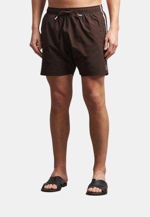 Uomo che indossa pantaloncini marrone scuro con coulisse e strisce laterali e sandali neri a ciabatta, in piedi davanti a uno sfondo semplice.