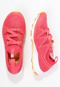 Klart rosa träningsskor med ett nätövre, gula detaljer, elastiska remmar för stängning och en vit, texturerad sula.