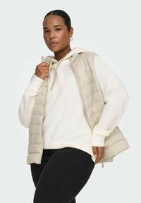 Dunvest i lys beige med quiltet design og glidelås, lagvis over en hvit hettegenser med gullhjerteemblem. Svarte jeans.