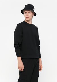 Sort sweatshirt med rund hals, lange ærmer og ribbede manchetter. Båret med sorte cargo-bukser og en sort bucket hat. Minimalistisk design.