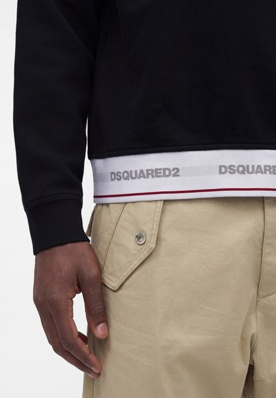 Gros plan d’une personne portant un sweat-shirt noir sur un pantalon beige avec une poche à rabat et une ceinture blanche visible étiquetée DSQUARED2.