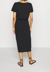 Robe midi en coton noir avec manches courtes et taille élastique, offrant une coupe décontractée et une texture lisse. Portée avec des sandales plates noires à lanières.