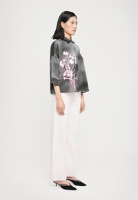 Camicia in seta oversize nelle tonalità di grigio con un motivo floreale rosa, colletto con bottoni e maniche a tre quarti, abbinata a pantaloni bianchi e tacchi neri.