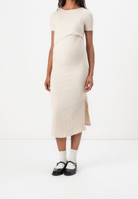 Robe de maternité en maille côtelée beige clair avec manches courtes, sur-couche courbée au niveau de la poitrine et fente latérale, portée avec des chaussures noires et des chaussettes blanches.